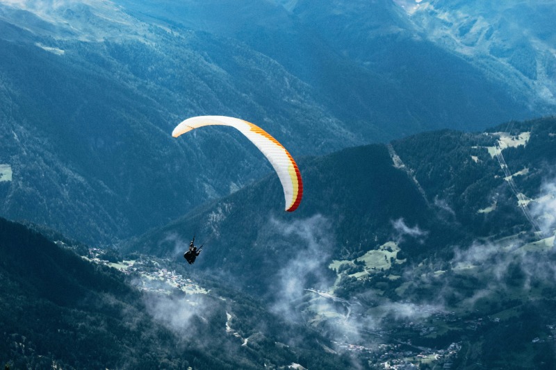 A person glides through the air on a paraglider, overlooking a stunning mountain range under a bright blue sky.