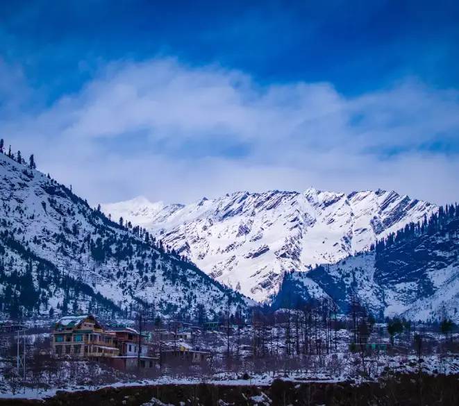 Snow-covered mountains with scattered trees and houses in a valley under a clear blue sky.