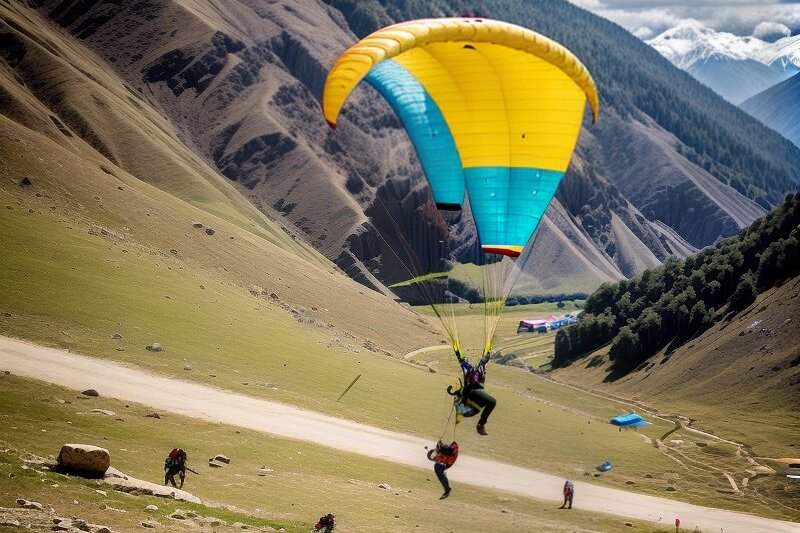 Two people paragliding with a multicolored parachute over a grassy valley, surrounded by mountains and trees. Other paragliders are visible on the ground.